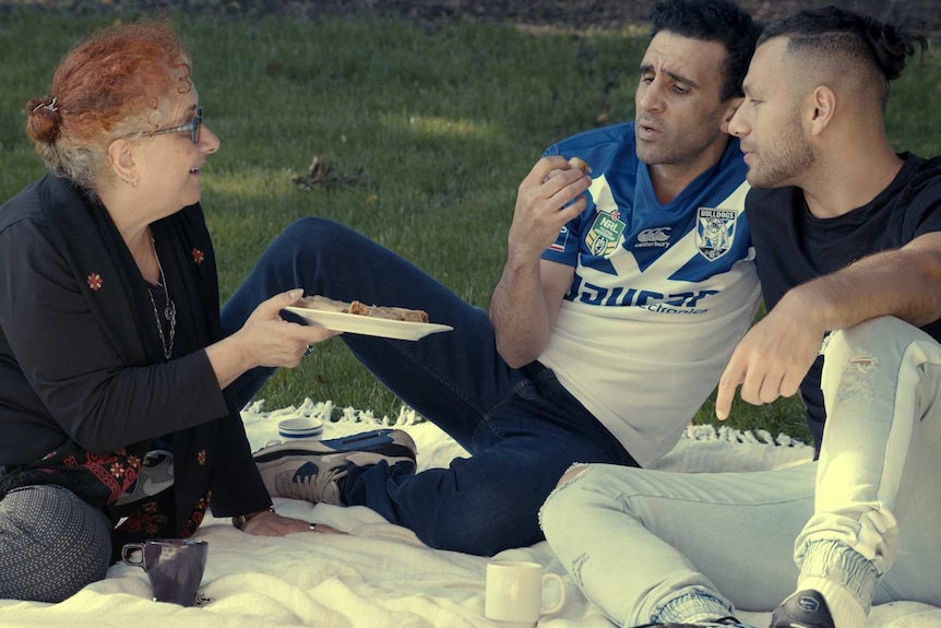 Two young men sit close together on white blanket on grass, while older woman sits on rug holding out a plate of food to them.