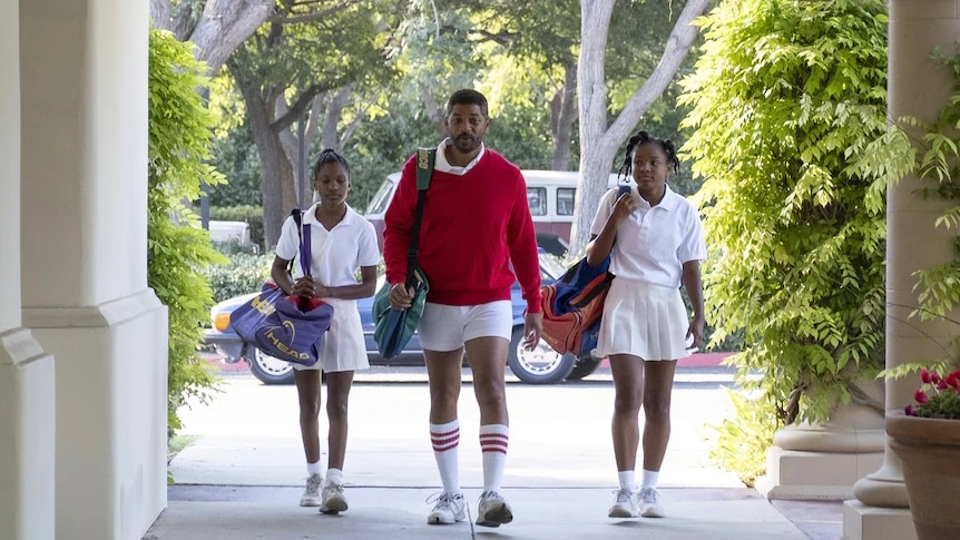 Will Smith wearing a red jumper and white shorts walking alongside two teenage girls holding tennis bags.