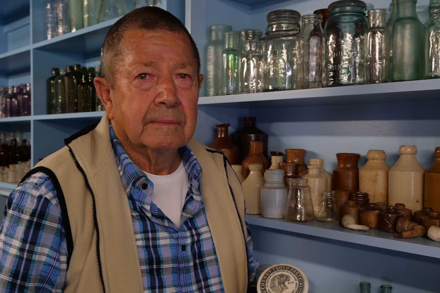 An older man standing in front of a collection of coloured glass bottles and other old artefacts.