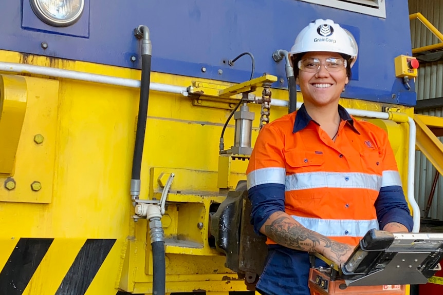 A maori woman wears a white hard hat and stands next to yellow equipment.