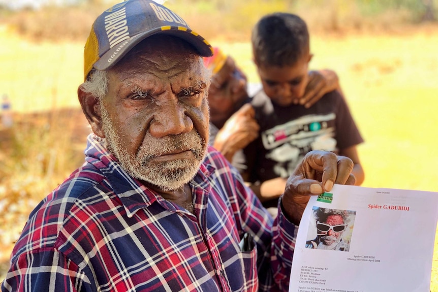 Image of an older indigenous man, wearing a checked shirt and baseball cap. He's holding a poster.