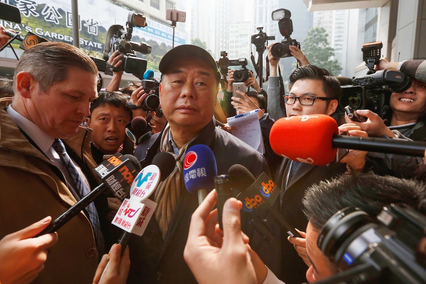 A man in a baseball cap surrounded by journalists holding microphones and cameras