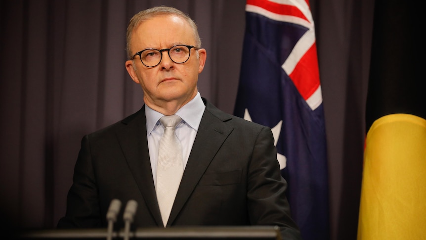 Anthony Albanese stands behind a lectern in a suit looking serious.