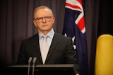 Anthony Albanese stands behind a lectern in a suit looking serious.