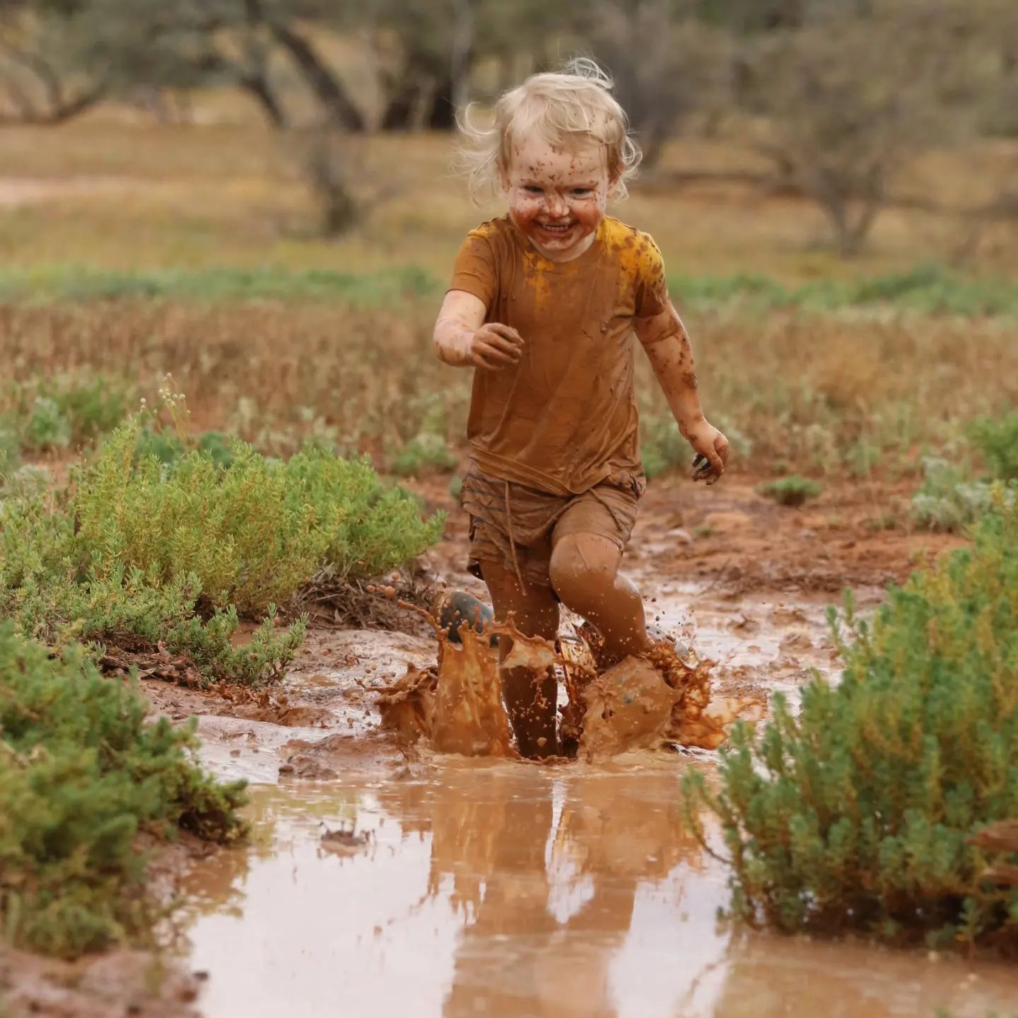 A young child runs in a long puddle of mud with a big grin, splashing muddy water