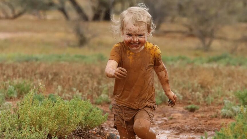 A young child runs in a long puddle of mud with a big grin, splashing muddy water.