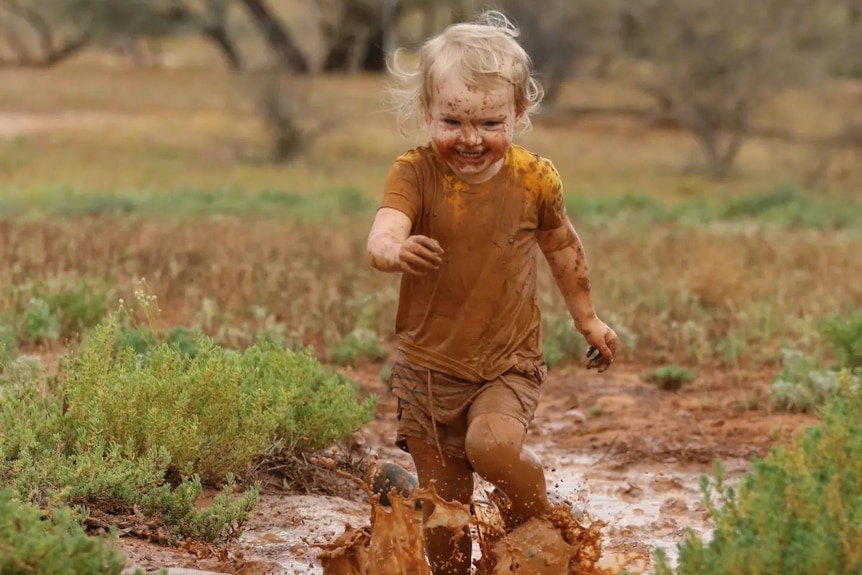 A young child runs in a long puddle of mud with a big grin, splashing muddy water