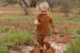 A young child runs in a long puddle of mud with a big grin, splashing muddy water.