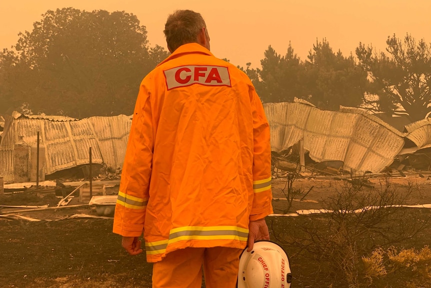 Steve Warrington surveying a destroyed house in Buchan wearing his orange CFA jacket.