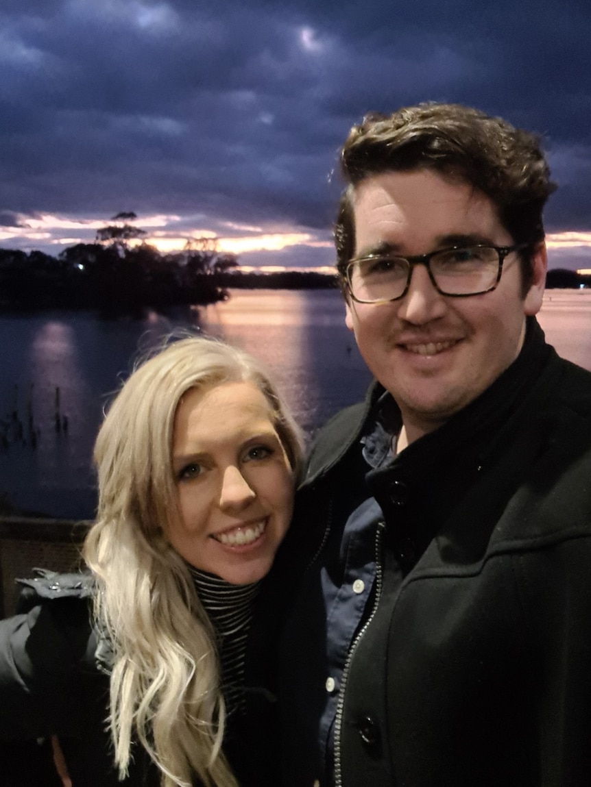 A smiling blonde woman and a dark-haired man cuddled up in front of a lake at sunset.