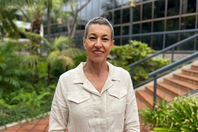 A woman stands in garden with grey short hair and beige shirt