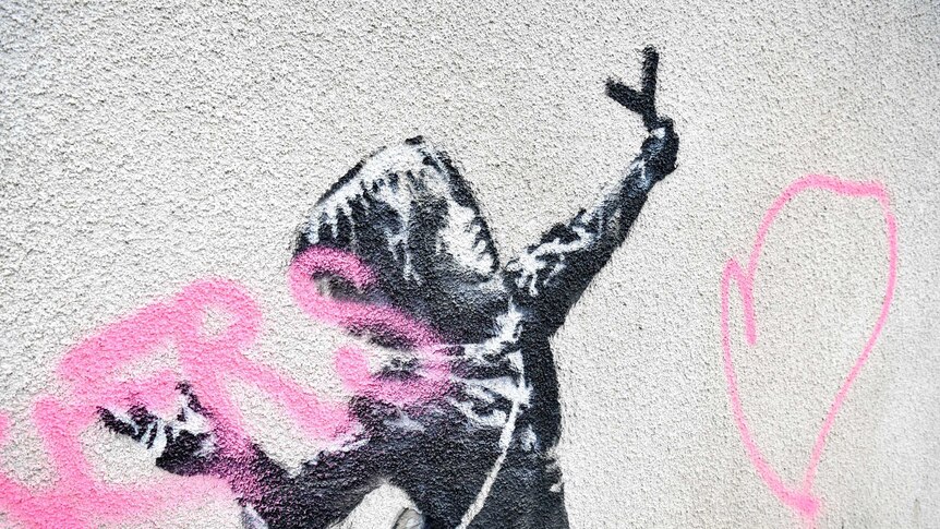 A view of a mural by Bansky which has been vandalised with pink spray paint.