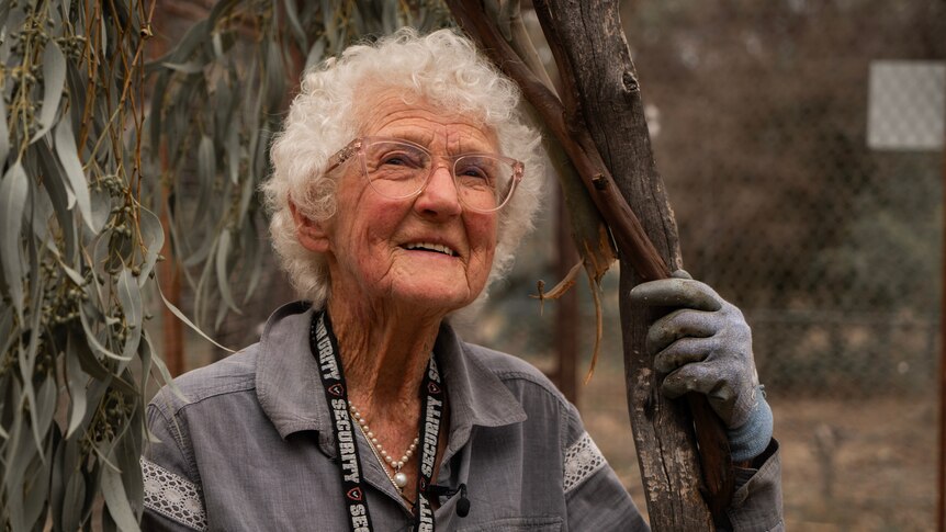 A woman, 90, looks upwards and holds onto a tree.