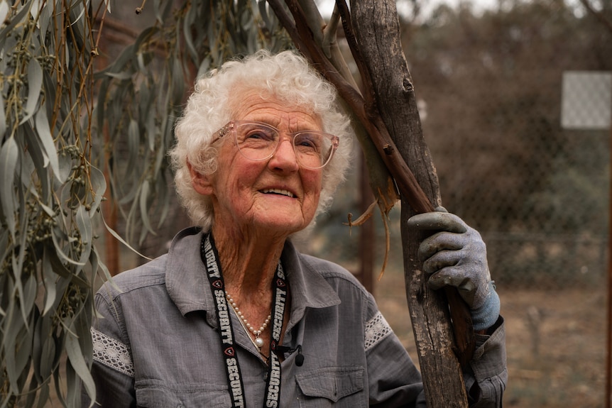A woman, 90, looks upwards and holds onto a tree.