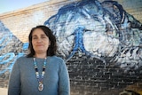 Lady standing in foreground with painted snake on wall in background, under a blue sky.