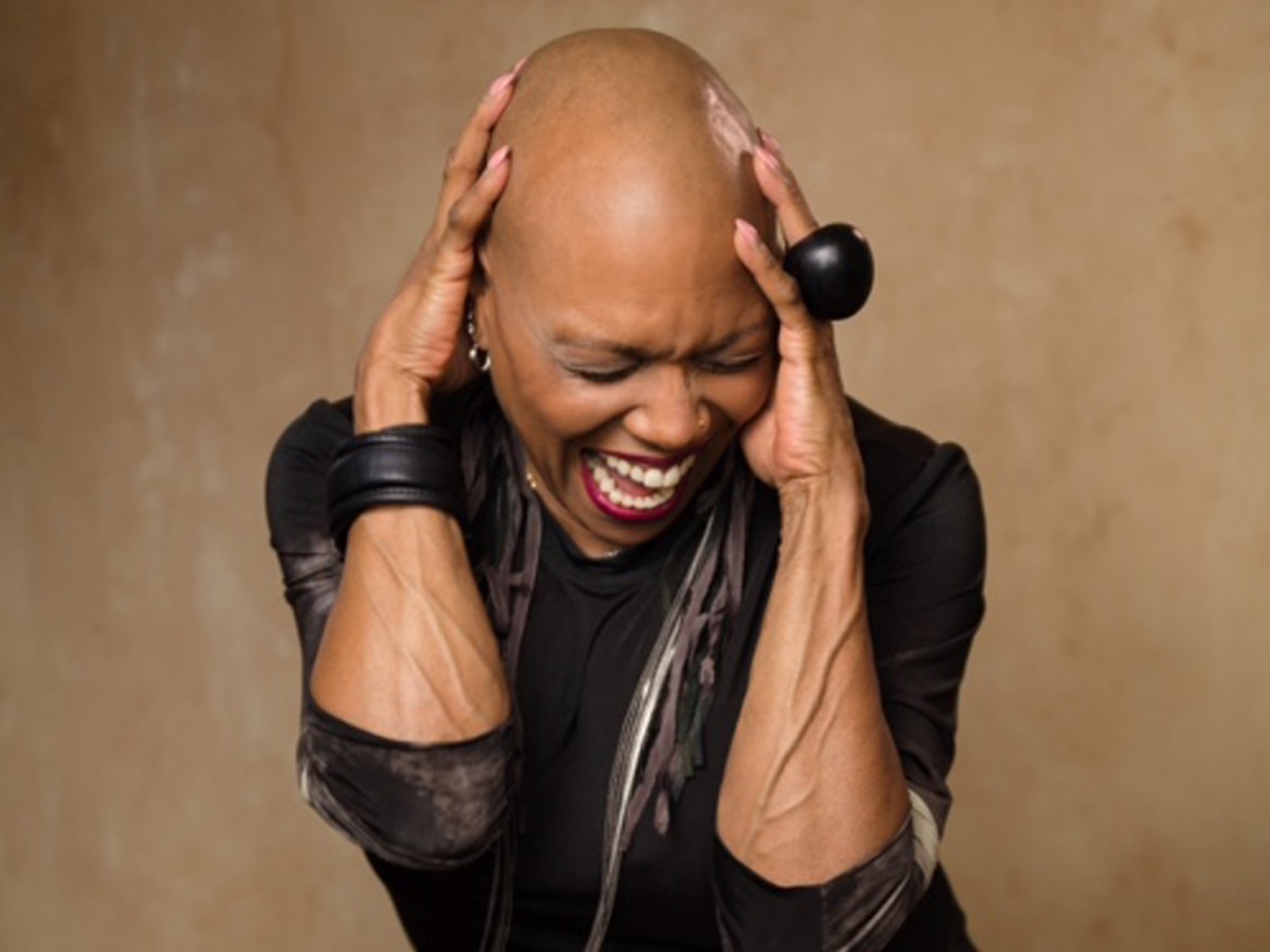Dee Dee Bridgewater leans forward with hands on head in a studio portrait. She wears black clothing and chunky jewellery.