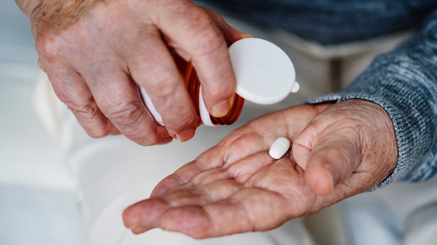 A pair of weathered, wrinkled male hands holds a plastic vessel in one hand and a tablet in the other