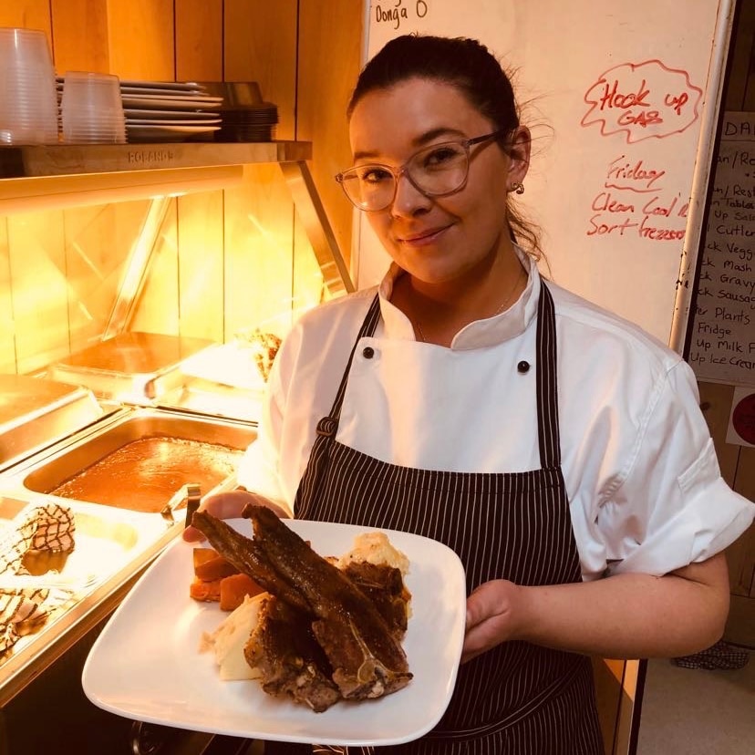 Woman in a chef's uniform stands in kitchen holding a plate of meat and mashed potato.