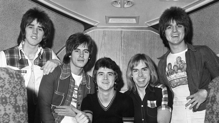 A black and white photo of five men in 70s style clothing on board a plane.