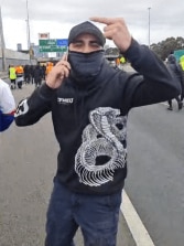 A man in a mask, wearing a jacket with a snake design, talks on the phone and makes an obscene gesture to the camera.