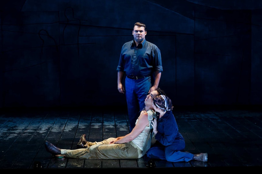 Three young actors on stage: a woman holds a man as another man watches over them