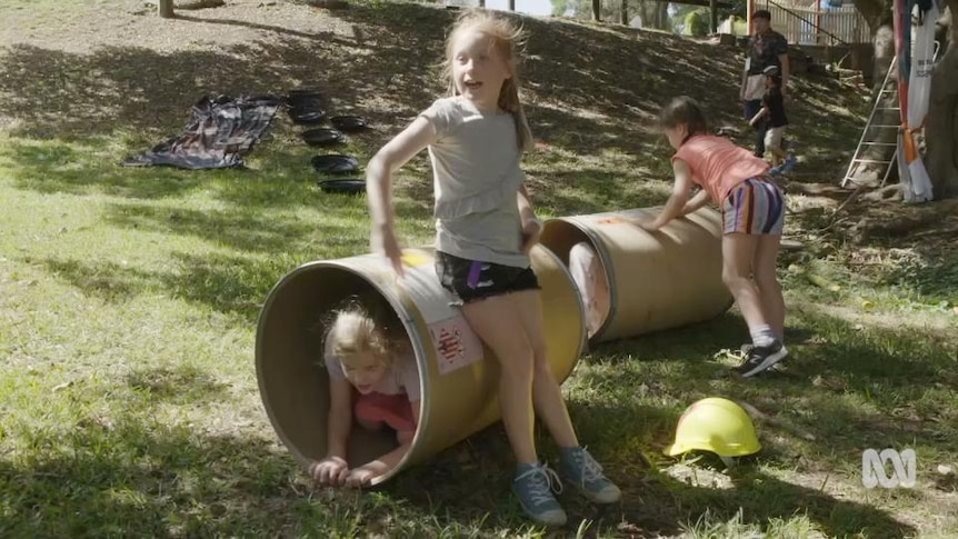 Children play on two large open drums on their sides