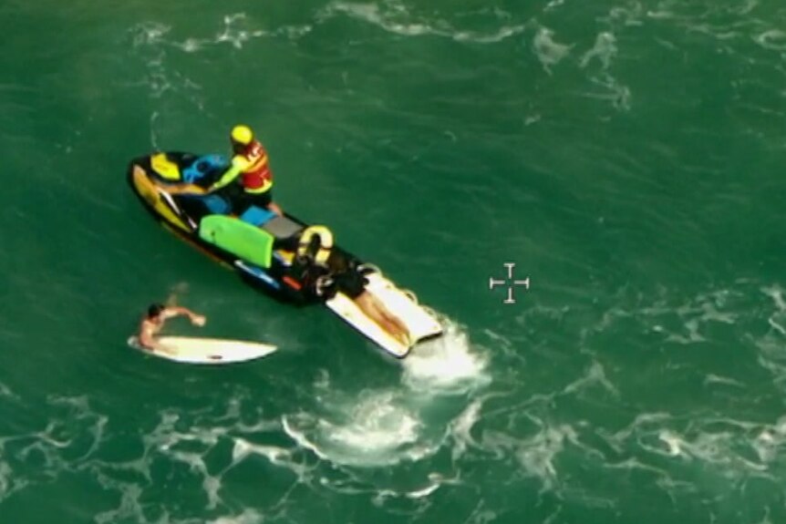 An aerial view of a man on a surfboard next to a surf lifesaving jet ski in the ocean.