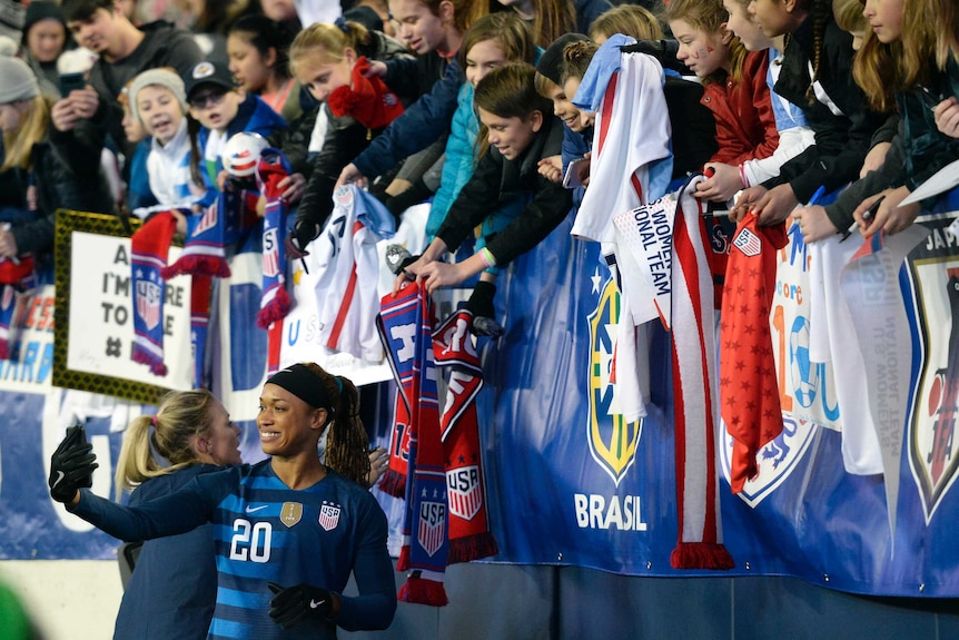 Jessica McDonald takes a selfie on the edge of the bleacher with fans holding flags and shirts smiling.