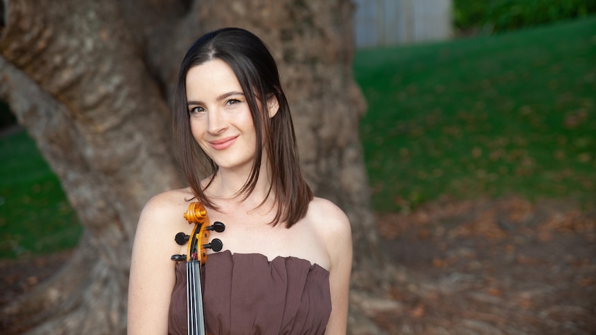 Violinist Amalia Hall with her violin standing in front of a tree
