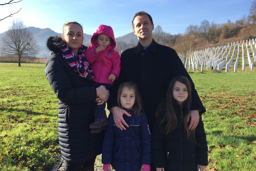 Avdic, wife and children standing in green field near Srebrenica memorial graves.