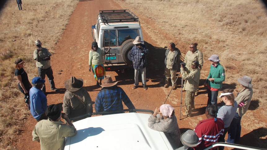 A group of people standing out in the countryside talking. Two cars are in the background