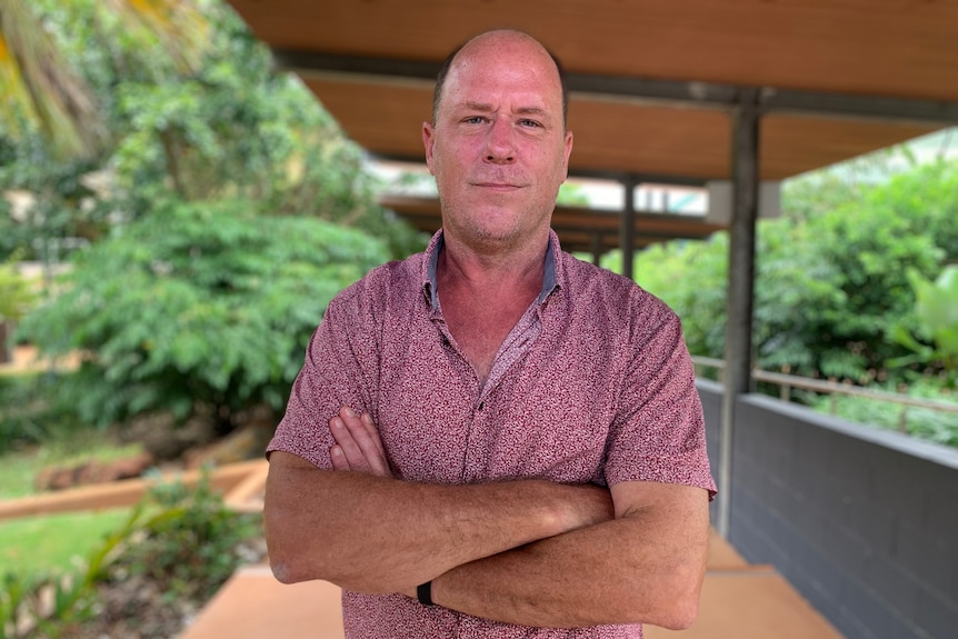 Dr Matt Stevens stands with his arms folded, looking towards the camera, with a walkway and trees in the background.