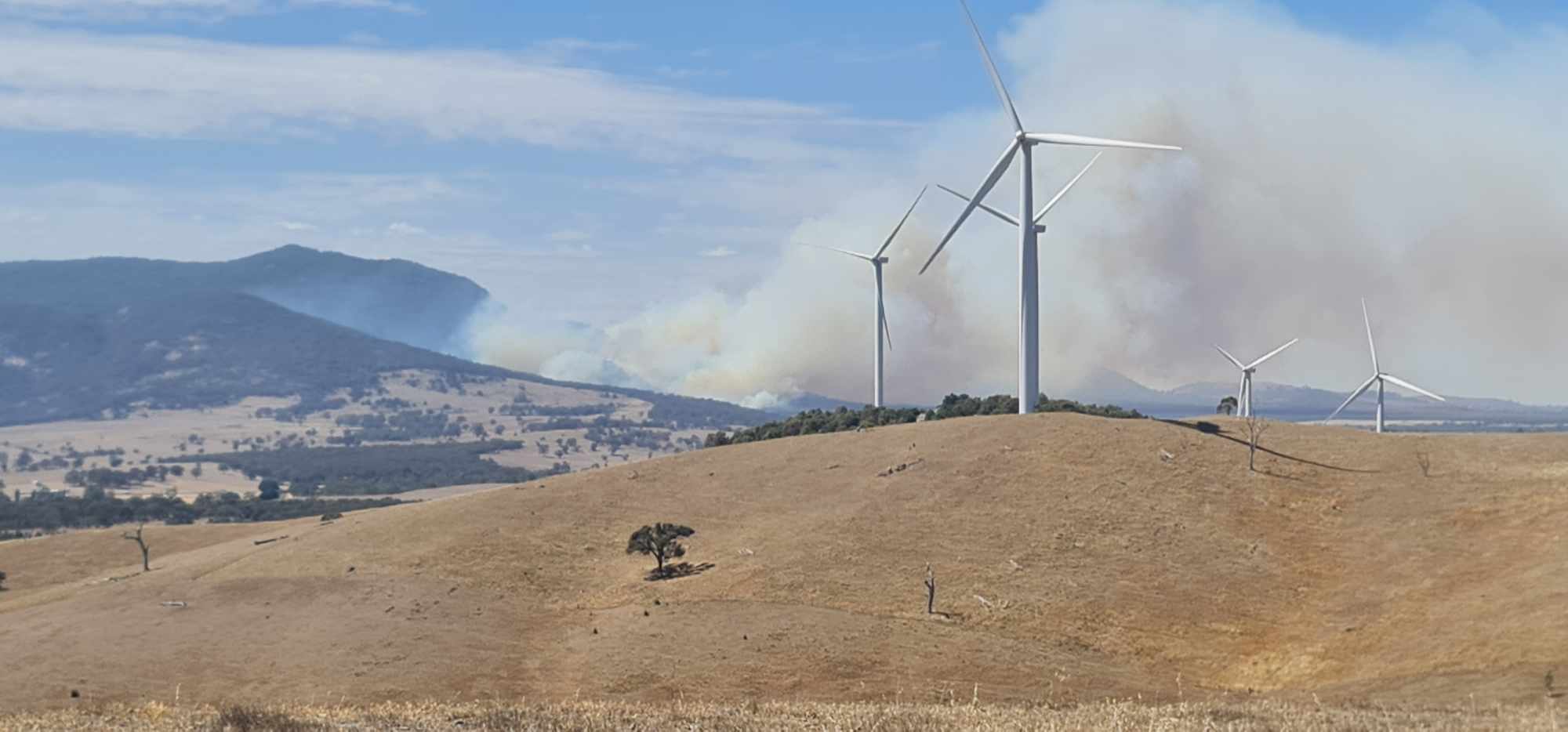 Smoke billowing behind a hill with wind turbines on it.