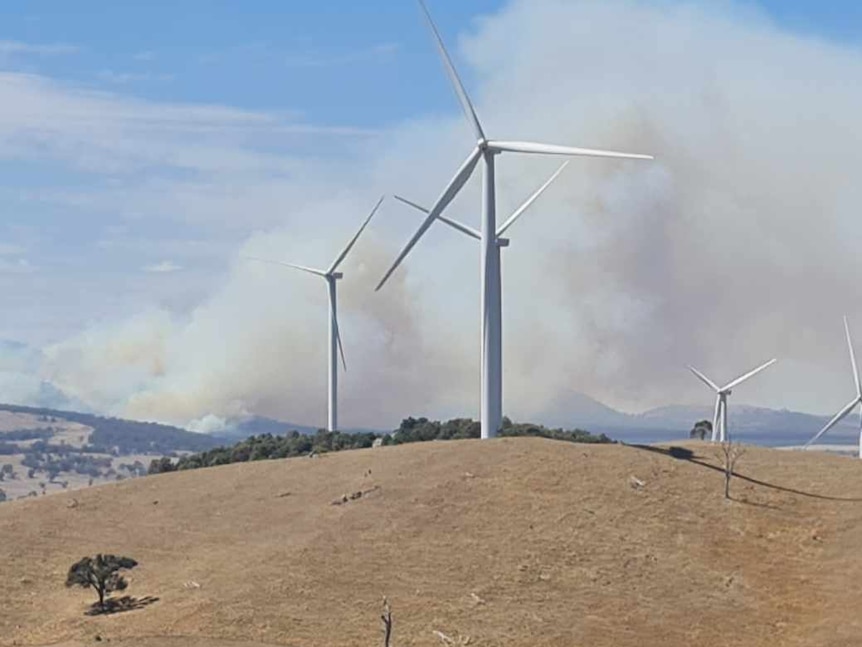 Smoke billowing behind a hill with wind turbines on it.