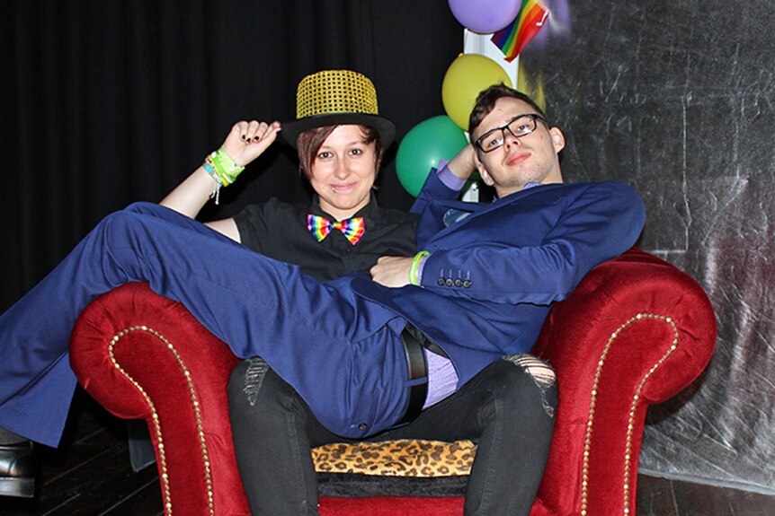 A young woman wearing a rainbow bow tie and top hat sits in a velvet chair with a young man in a blue suit seated on her lap