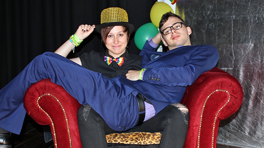 A young woman wearing a rainbow bow tie and top hat sits in a velvet chair with a young man in a blue suit seated on her lap