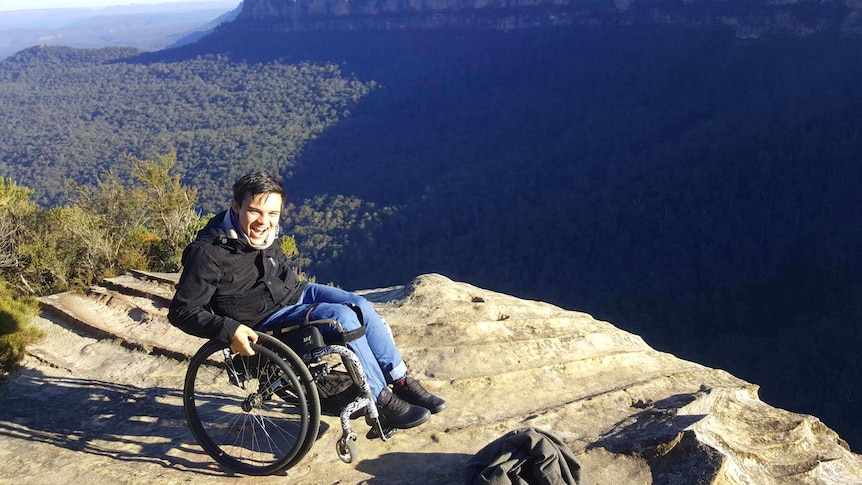 A laughing young man in a wheelchair does a wheelie on the edge of a cliff.