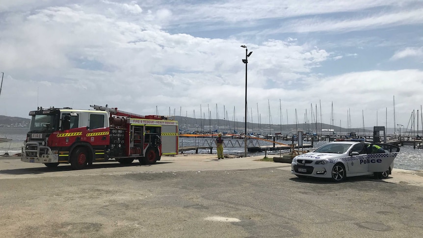 A wide shot of a fire truck and a police car parked along the water in front of boats at Royal Princess Sailing Club in Albany.