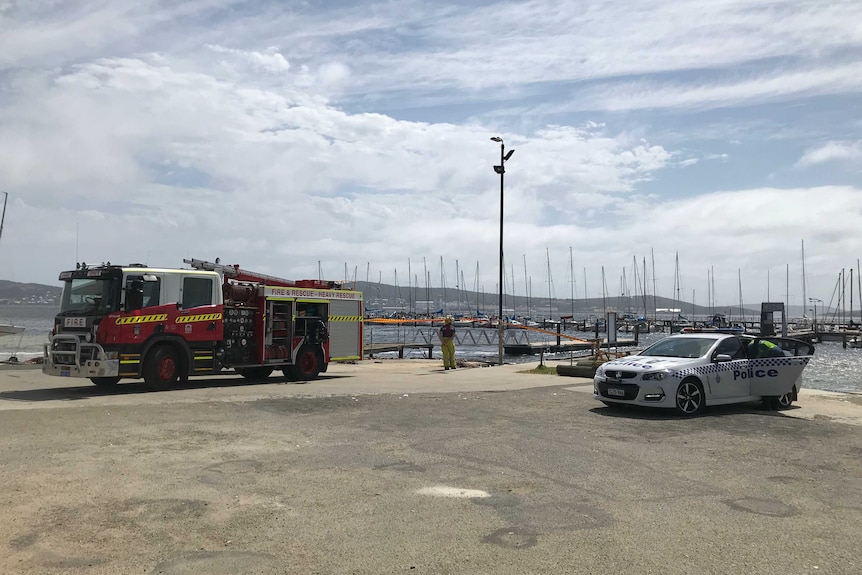 A wide shot of a fire truck and a police car parked along the water in front of boats at Royal Princess Sailing Club in Albany.