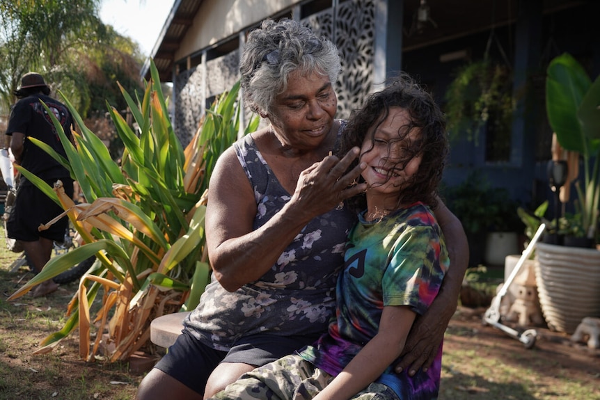 Indigenous grandma sits on a chair in her backyard and hugs her grandson 