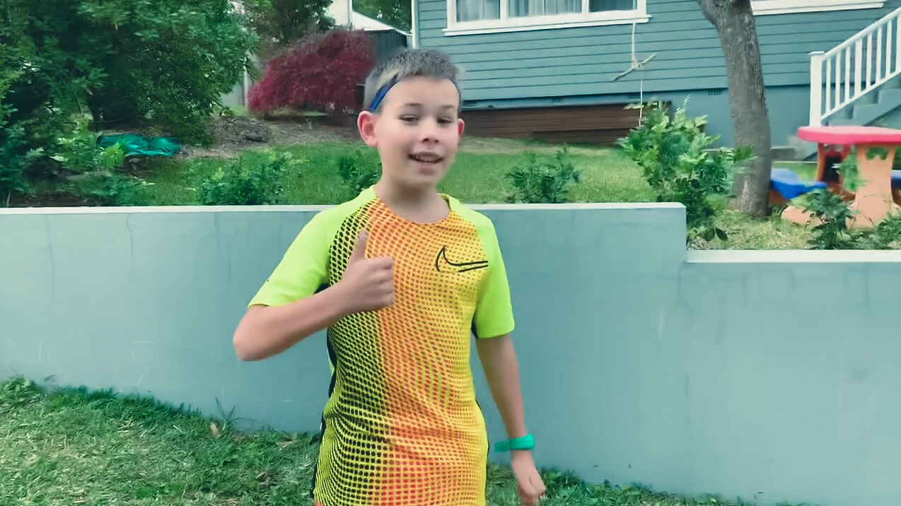A boy wearing a headband and sporting clothes stands holding a thumb up while standing on the sidewalk in front of a home.