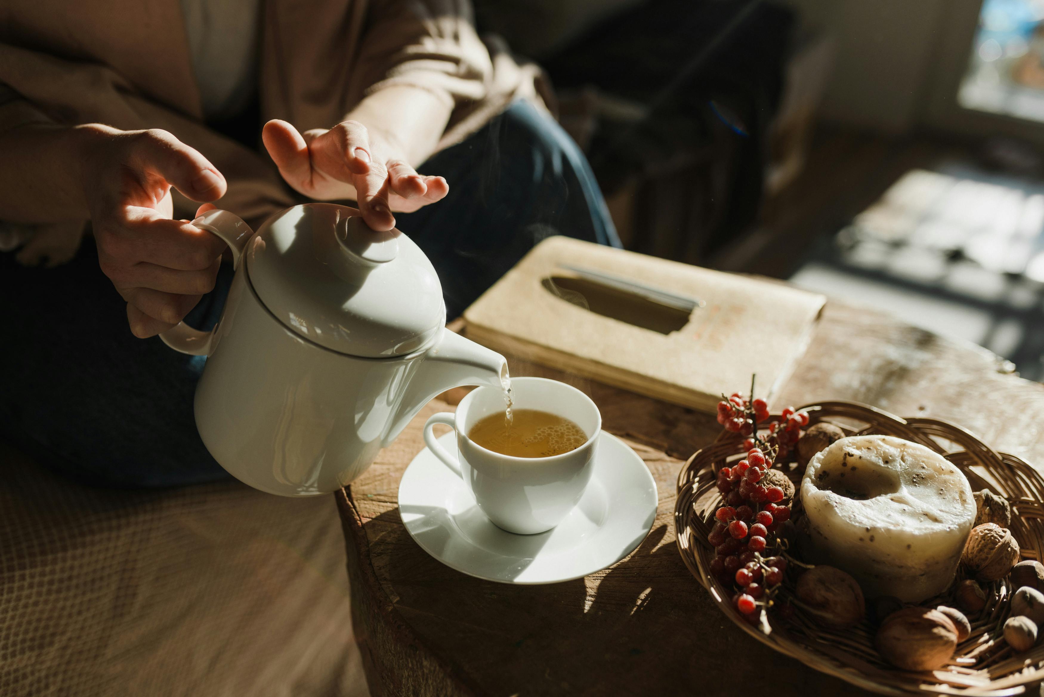 a cup of tea being poured by a teapot in front of a window with light streaming in. A wheel of cheese is nearby with grapes.