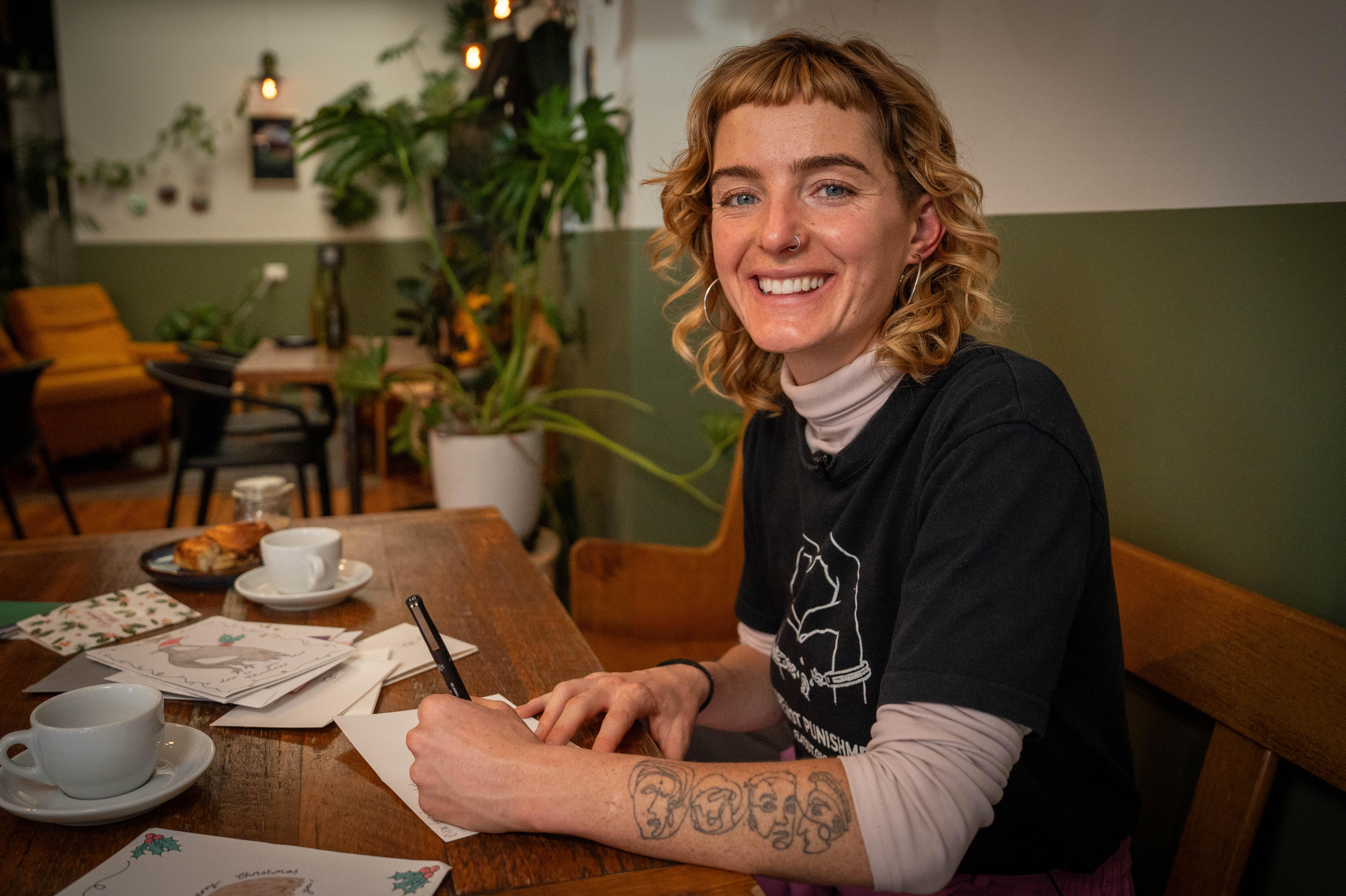 Una mujer sonríe a la cámara mientras escribe una tarjeta en un café. Sobre la mesa hay varias cartas y tazas de café.