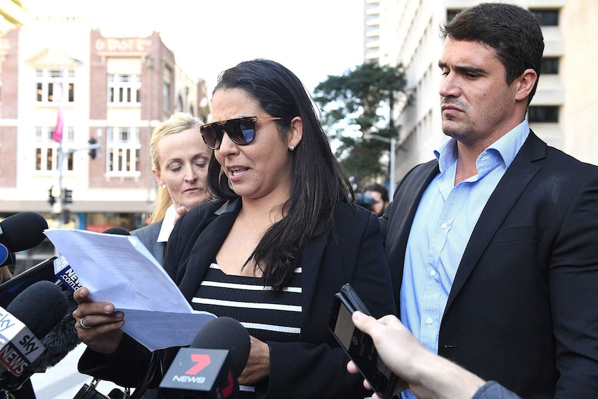 A woman in sunglasses delivers a statement to media microphones, a man standing at her side