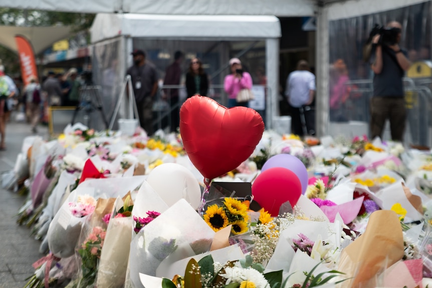 The camera focuses on a red heart balloon floating above a large group of flowers on a street.