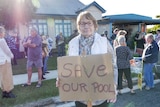 An elderly woman holds a sign in front of a group of protesters that reads "save our pool".