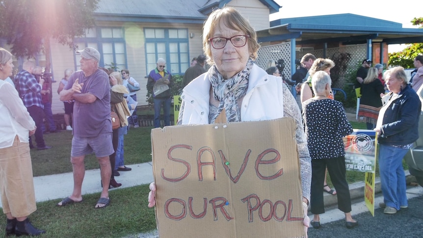 An elderly woman holds a sign in front of a group of protesters that reads "save our pool".