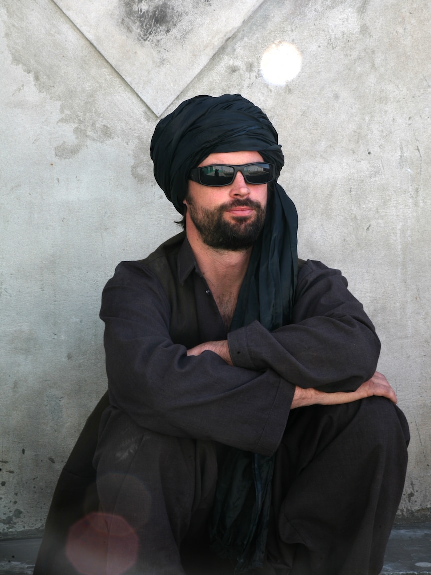 A bearded man sits in black sunglasses, a black headscarf and black outfit.