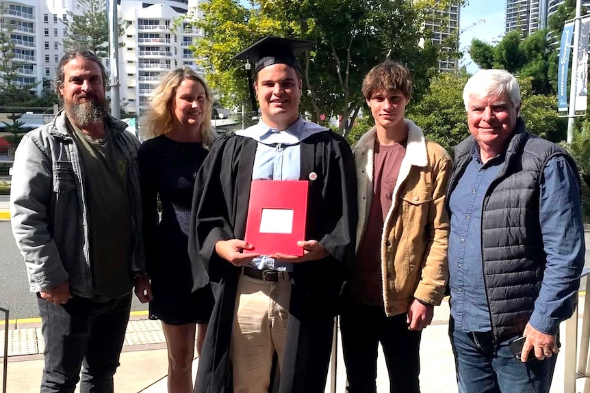 A man with grey hair, one with long beard, one woman, a boy stand behind a man wearing graduation gown, holding red folder.