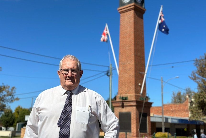 A man in a white shirt and tie stands in front of a brick clock tower memorial with two flags on it.